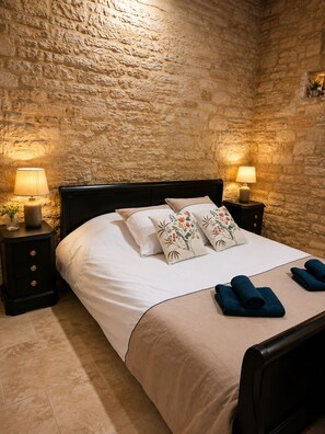 A bed with a white mattress, two decorative pillows, a beige bedspread, and two rolled towels. There are two bedside tables with lamps, and the room has a stone wall. - The "White Lady" Bed and Breakfast Suite (Borrèze)