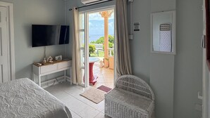 A hotel room with a bed, a desk, a television, an air conditioning unit, a wicker chair, and a view of the outdoors. - Belle Escape - Blue Room (Castries)