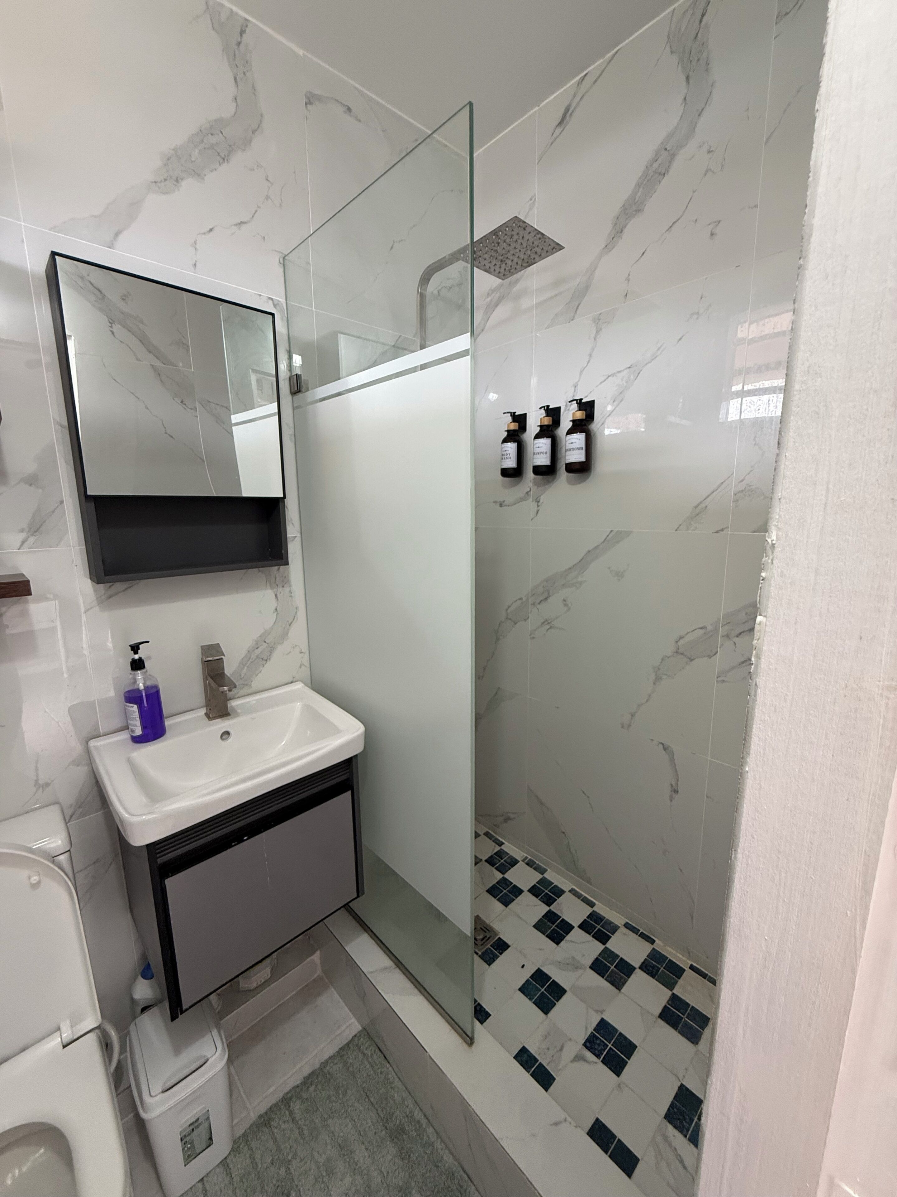 Un baño moderno con ducha revestida de mármol, cabina de ducha con paredes de vidrio, lavamanos blanco con espejo y un pequeño cubo de basura.