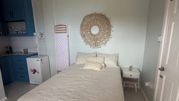 A bedroom with a bed, a bedside table, a wall decoration, and a kitchen area with cabinets and a refrigerator. - Belle Escape - Blue Room (Castries)