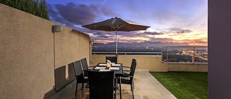 An outdoor patio with a table and chairs, a large umbrella, and a view of a cityscape at dusk.