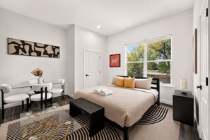 A modern bedroom with a large bed, a desk, and a chair. There is a window with a view of trees. - Settle into a modern, private upper-floor studio designed to make an extended Dallas stay feel easy and comfortable. (Dallas)