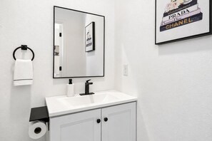 A bathroom with a white sink, black faucet, and a mirror above it. There is a framed picture of luxury brands on the wall. - Settle into a modern, private upper-floor studio designed to make an extended Dallas stay feel easy and comfortable. (Dallas)