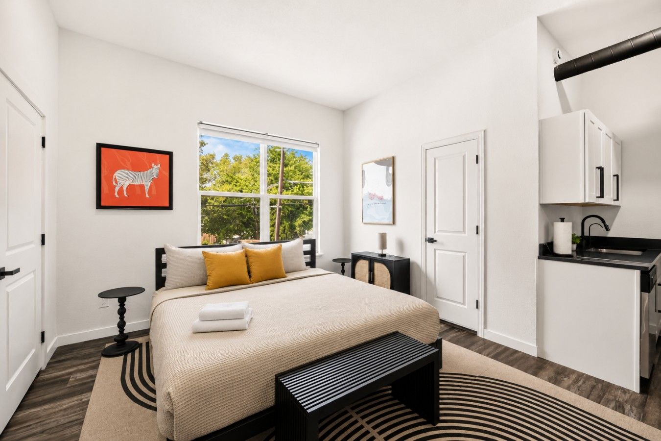 Quarto moderno com cama, janela, escrivaninha e pequeno espaço de cozinha.