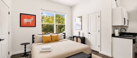 Quarto moderno com cama, janela, escrivaninha e pequeno espaço de cozinha.