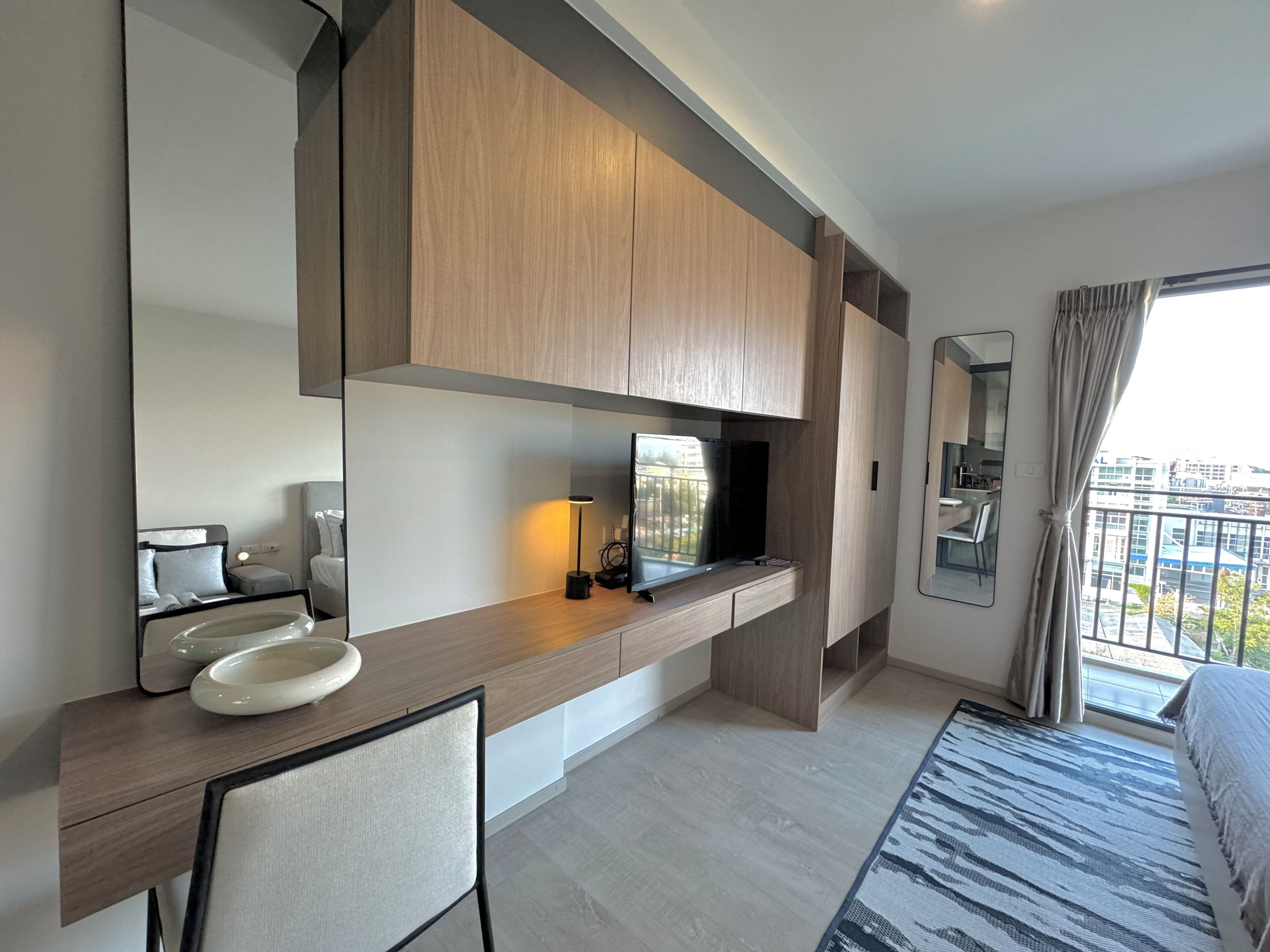A modern hotel room with a wooden vanity, a mirror, a bed, and a view of the city.