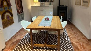 Een eetgedeelte met een houten tafel en stoelen, een kamerkleed met een patroon, een keuken op de achtergrond en een open haard.