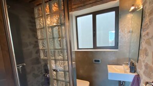 Een badkamer met een douchecabine van glas, een wit toilet, een wastafel met spiegel en een raam boven de wastafel.