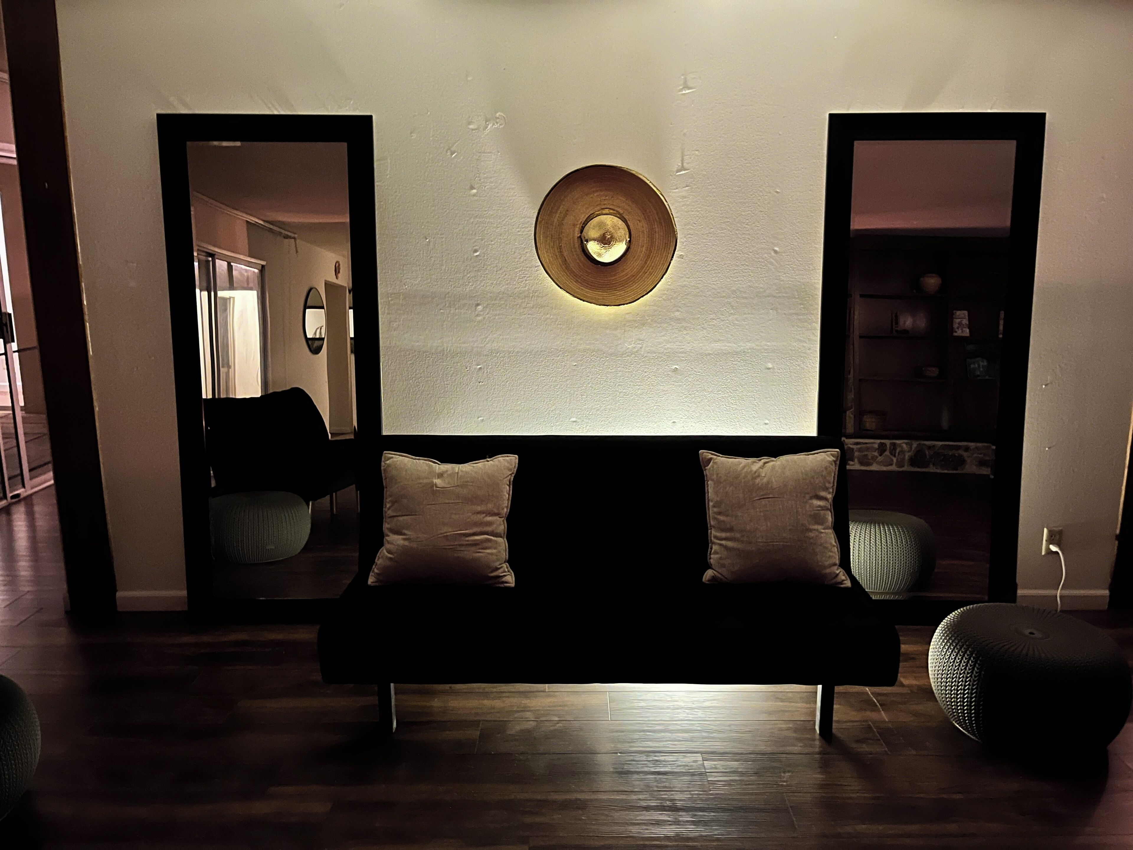 A modern living room with a dark-colored sofa, two cushions, a round wall-mounted light fixture, and a wooden floor.