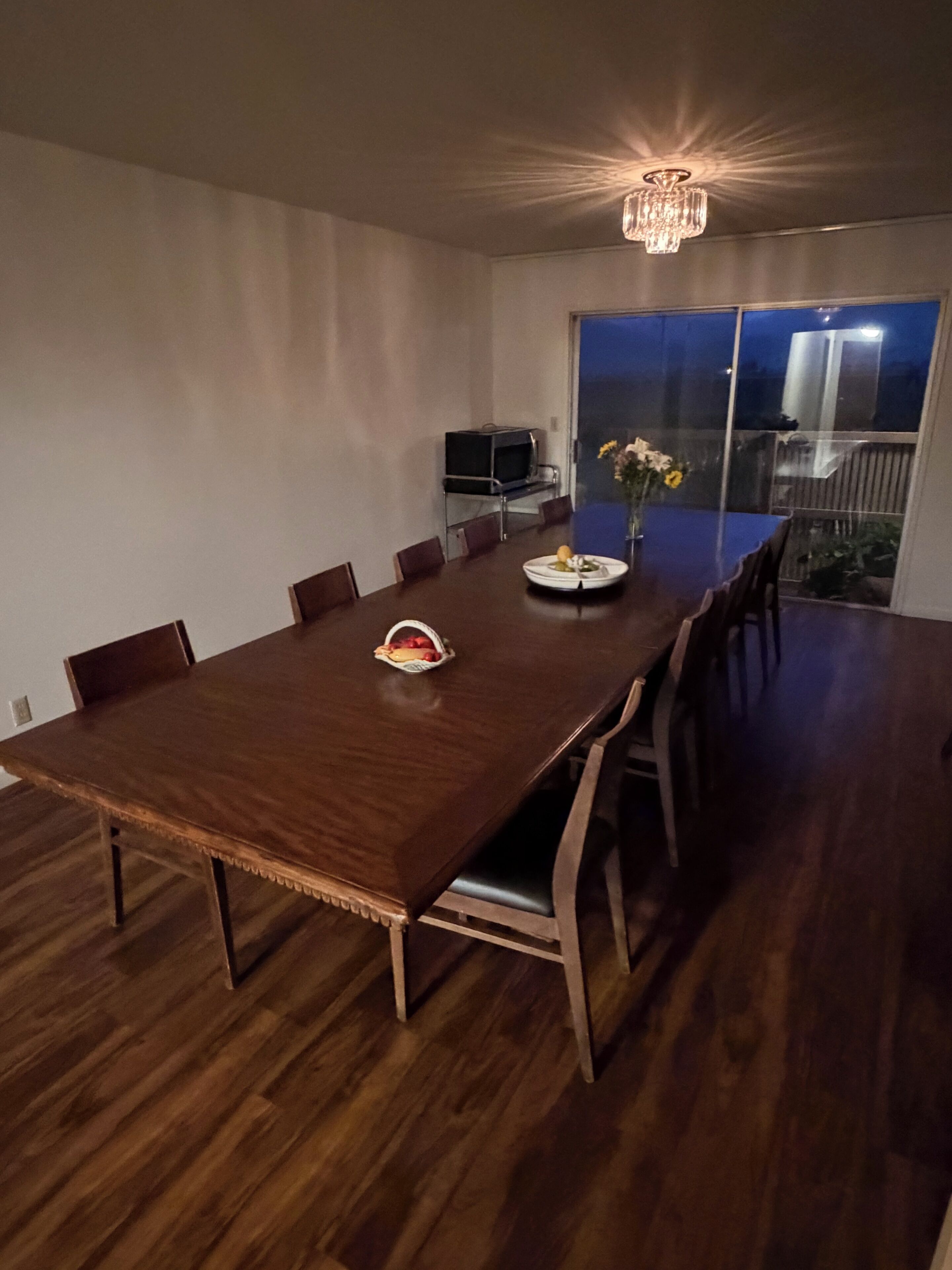 Un coin repas avec une grande table en bois, des chaises, un lustre et un téléviseur posé sur un meuble.