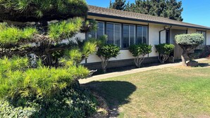 Una casa con un césped cuidado, un gran pino y un árbol más pequeño de copa redondeada.