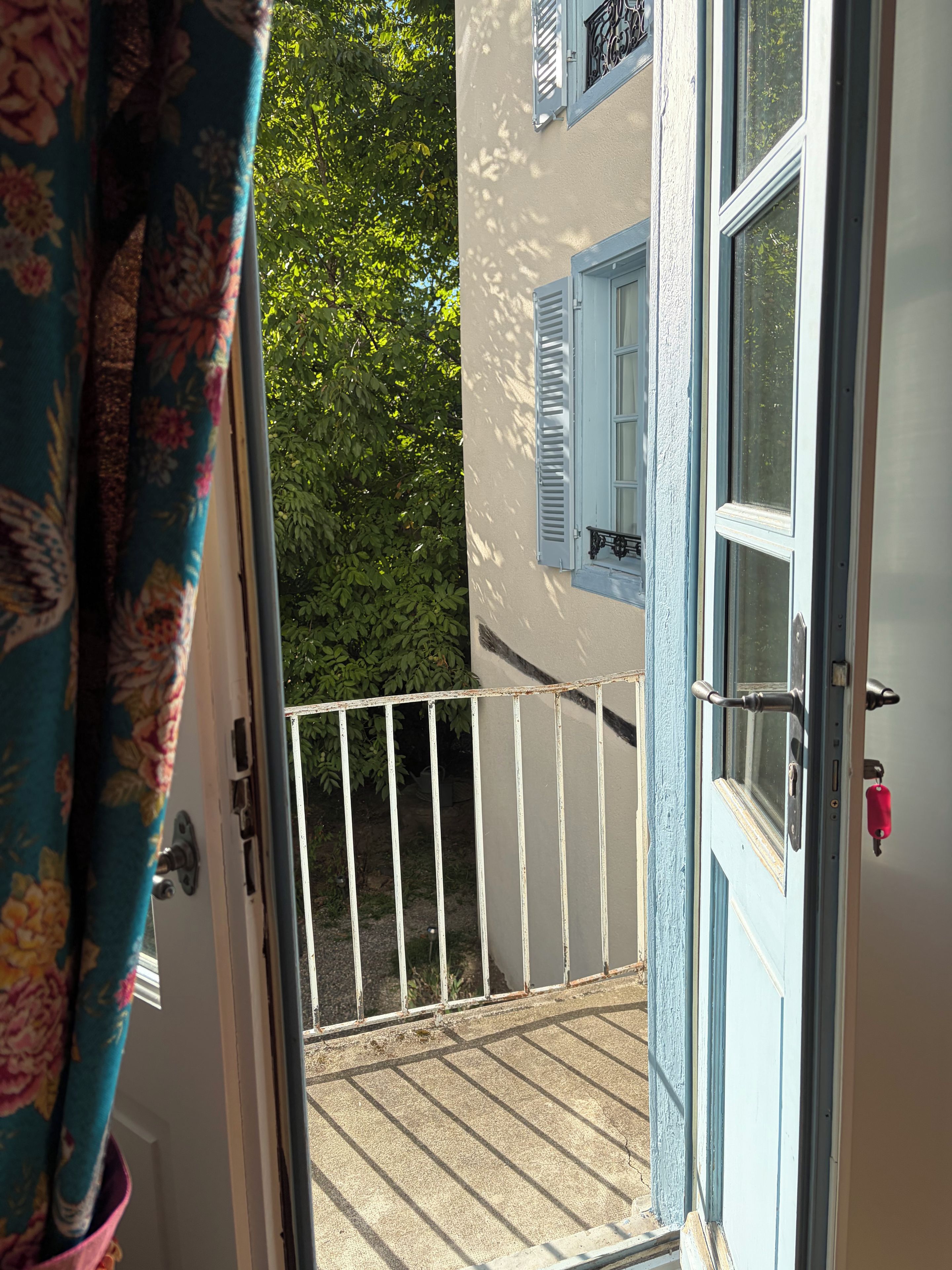 An open door to a balcony with a white railing, a blue door with a red handle, and a window with blue shutters.