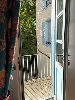 An open door to a balcony with a white railing, a blue door with a red handle, and a window with blue shutters.