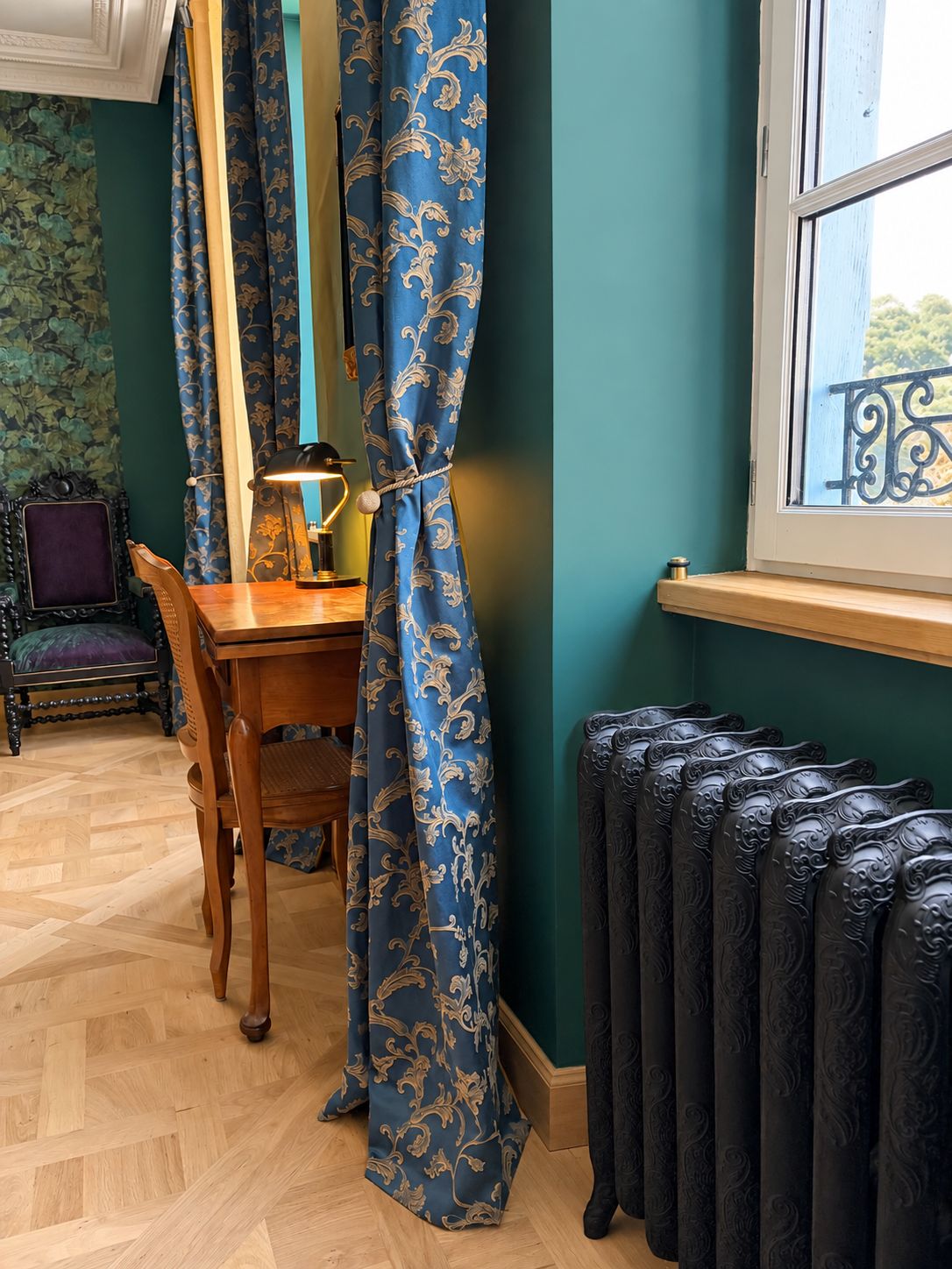 A room with a wooden table, a chair, a lamp, a radiator, and a window with a view.