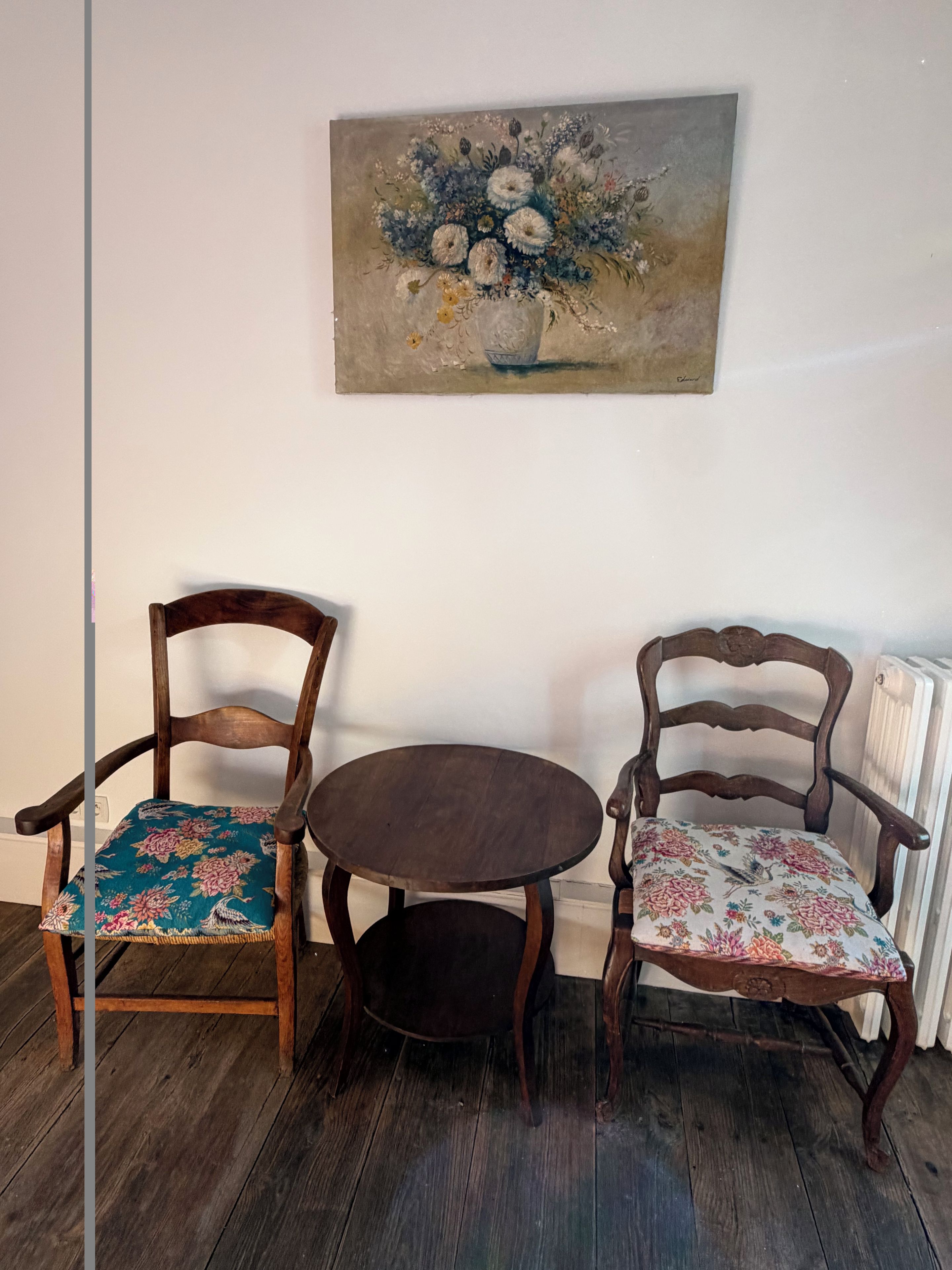 Een kamer met twee houten stoelen met bloemenmotieven, een klein rond tafeltje en een bloemschilderij aan de muur.