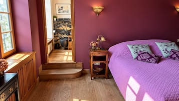 Une chambre avec une couvre-lit violette, une table de chevet en bois avec une lampe, et une vue sur un couloir doté d’un plancher en bois et d’une porte.