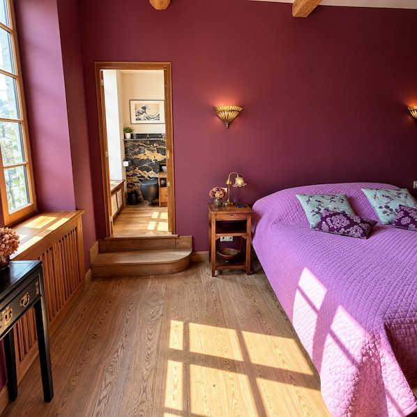 A bedroom with a purple bedspread, a wooden nightstand with a lamp, and a view of a hallway with a wooden floor and a door.