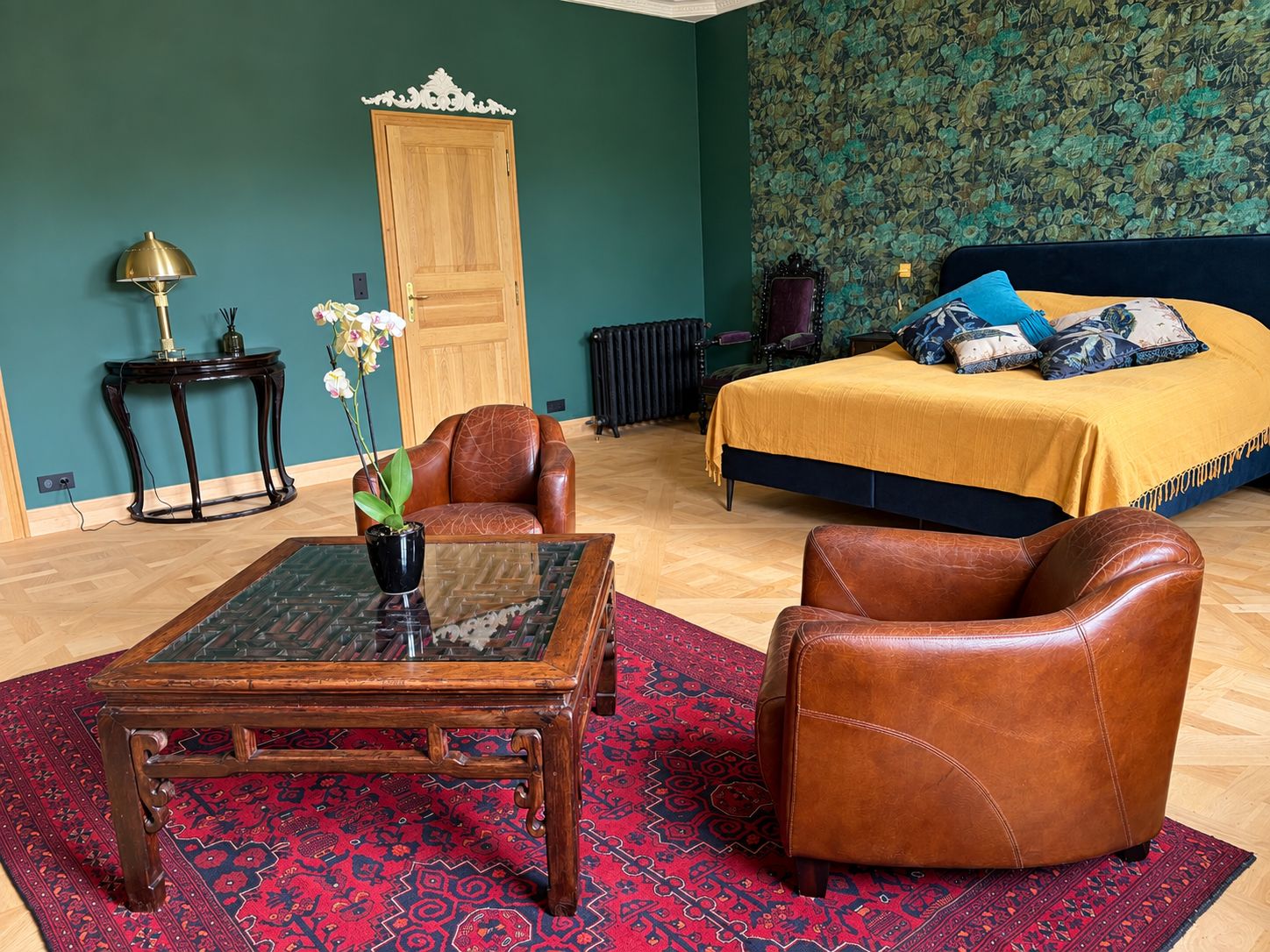 A bedroom with a bed, two leather chairs, a glass-top table, a potted plant, and a radiator.