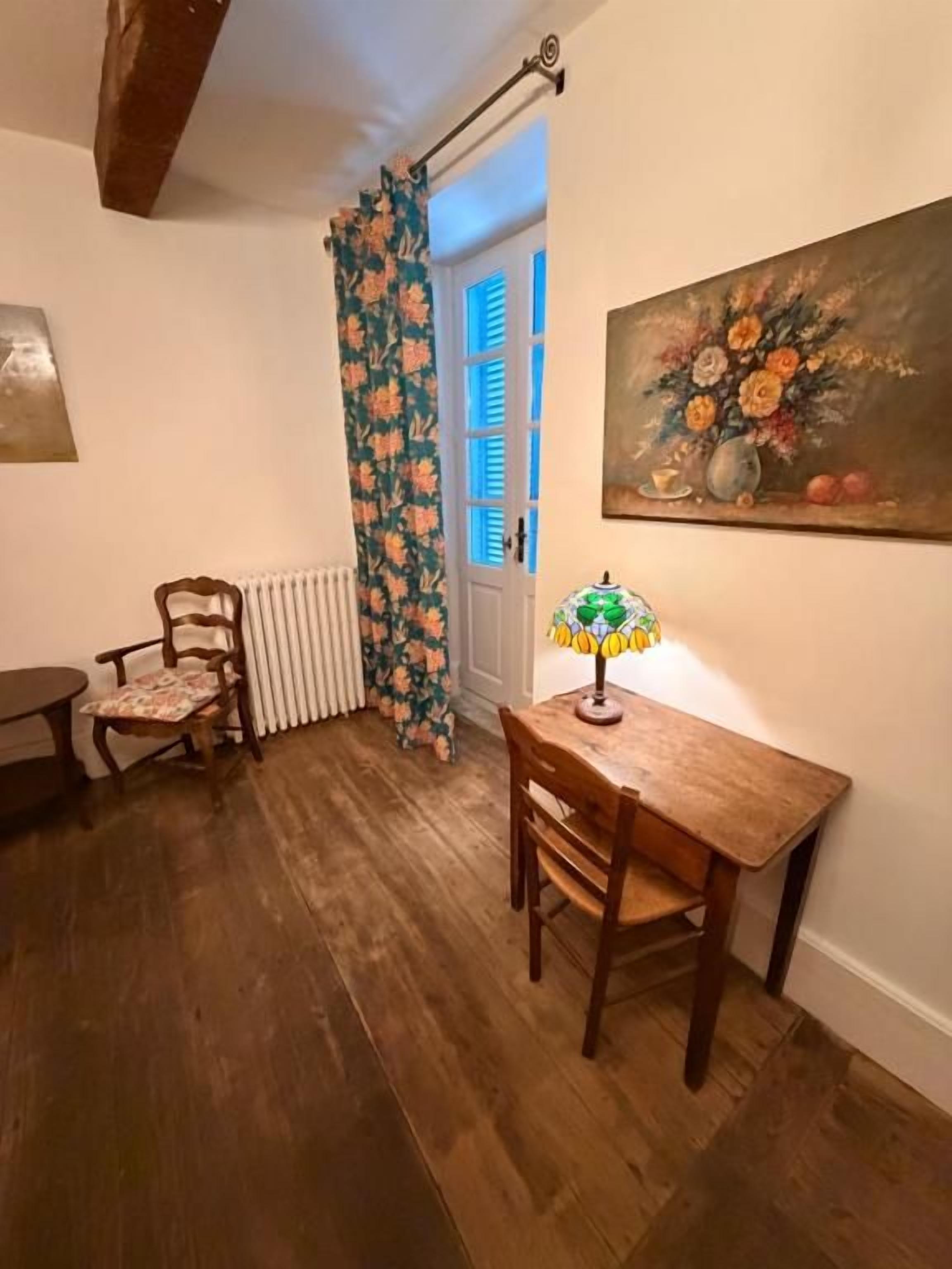 Une pièce avec un plancher en bois, une table avec une lampe, une chaise, un radiateur et une porte avec des volets bleus.