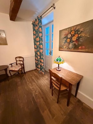 A room with a wooden floor, a table with a lamp, a chair, a radiator, and a door with blue shutters.