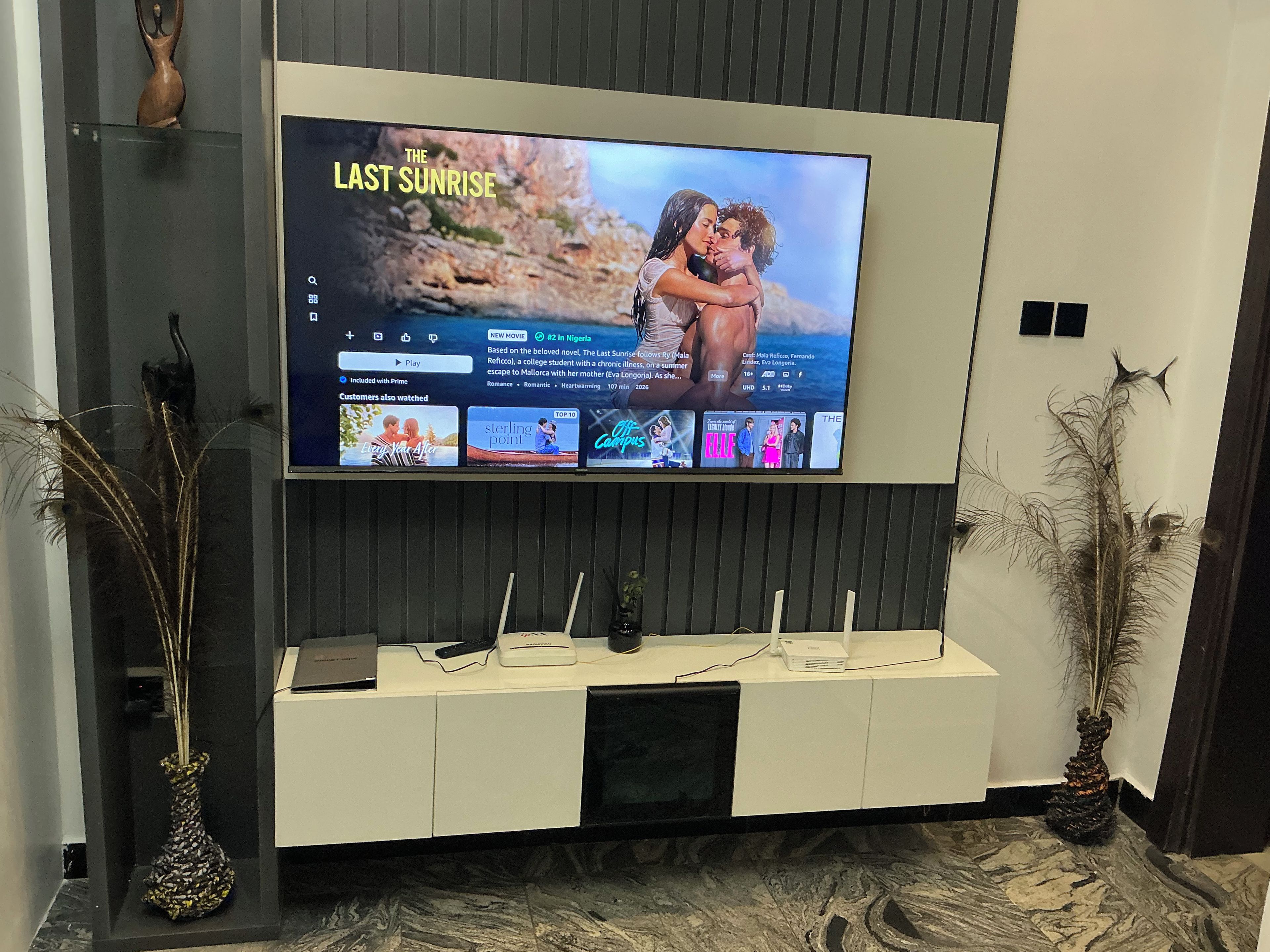 A flat-screen TV mounted on a wall above a white entertainment unit with a remote control, a speaker, and decorative items.