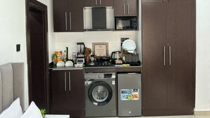 A compact kitchen with dark cabinets, a microwave, a coffee maker, and a washing machine. - Lexrach Luxury Apartments (Abuja)