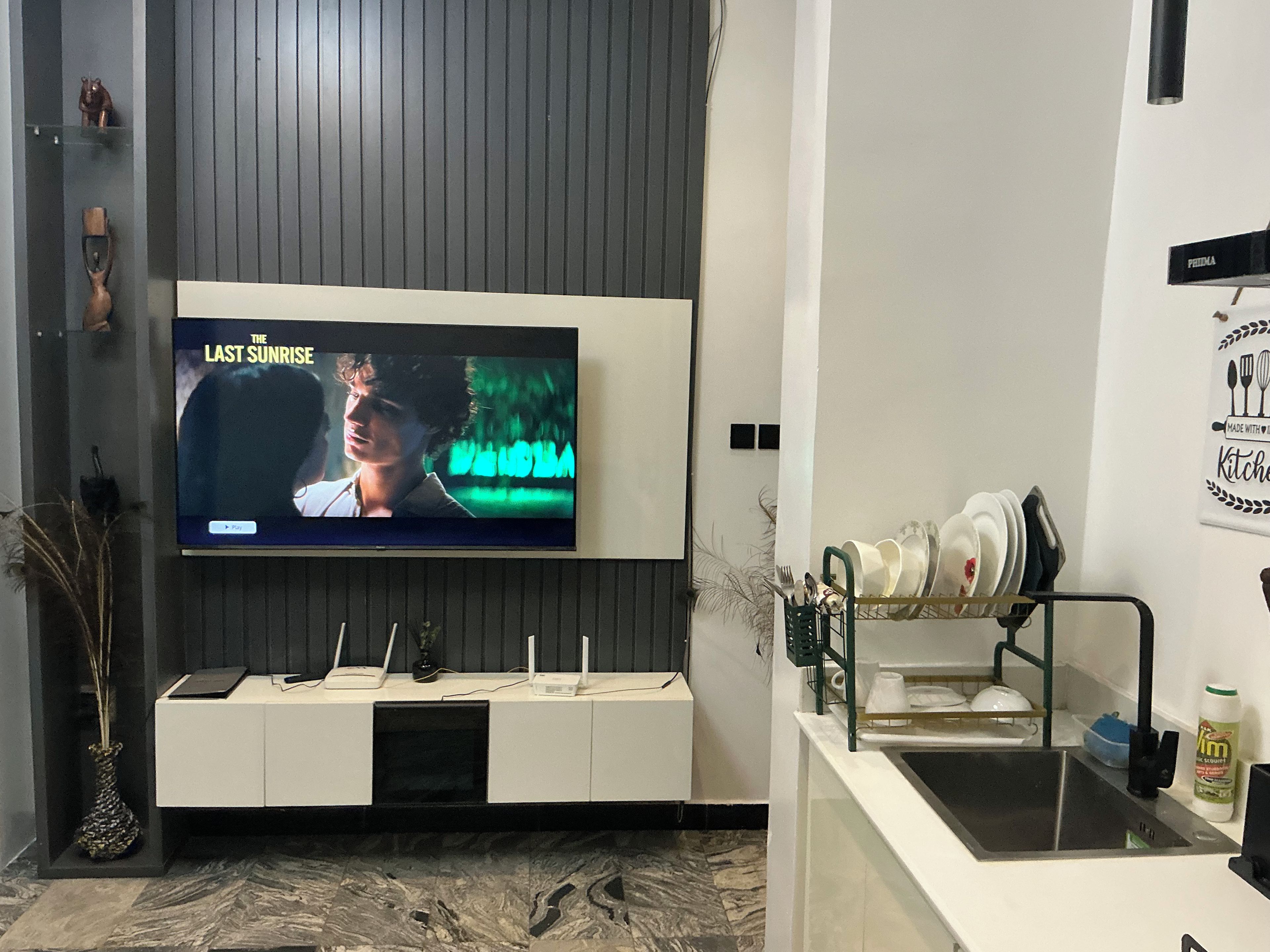 A modern living room with a flat-screen TV mounted on a wall, a white entertainment unit below, and a kitchen area with a sink and dish rack.