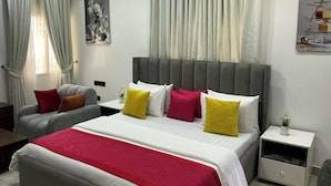 A modern hotel room with a large bed, a grey upholstered armchair, a bedside table with a lamp, and a painting on the wall. - Lexrach Luxury Apartments (Abuja)