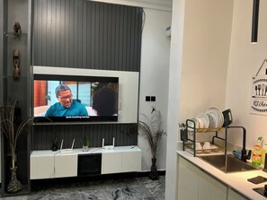 A modern living room with a flat-screen TV mounted on a dark wall, a white console table with decorative items, and a kitchen area with a sink and dish rack. - Lexrach Luxury Apartments (Abuja)