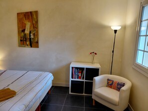 Ein Schlafzimmer mit einem Bett, einem weißen Sessel, einem Bücherregal, einer Stehlampe und einem Gemälde an der Wand.