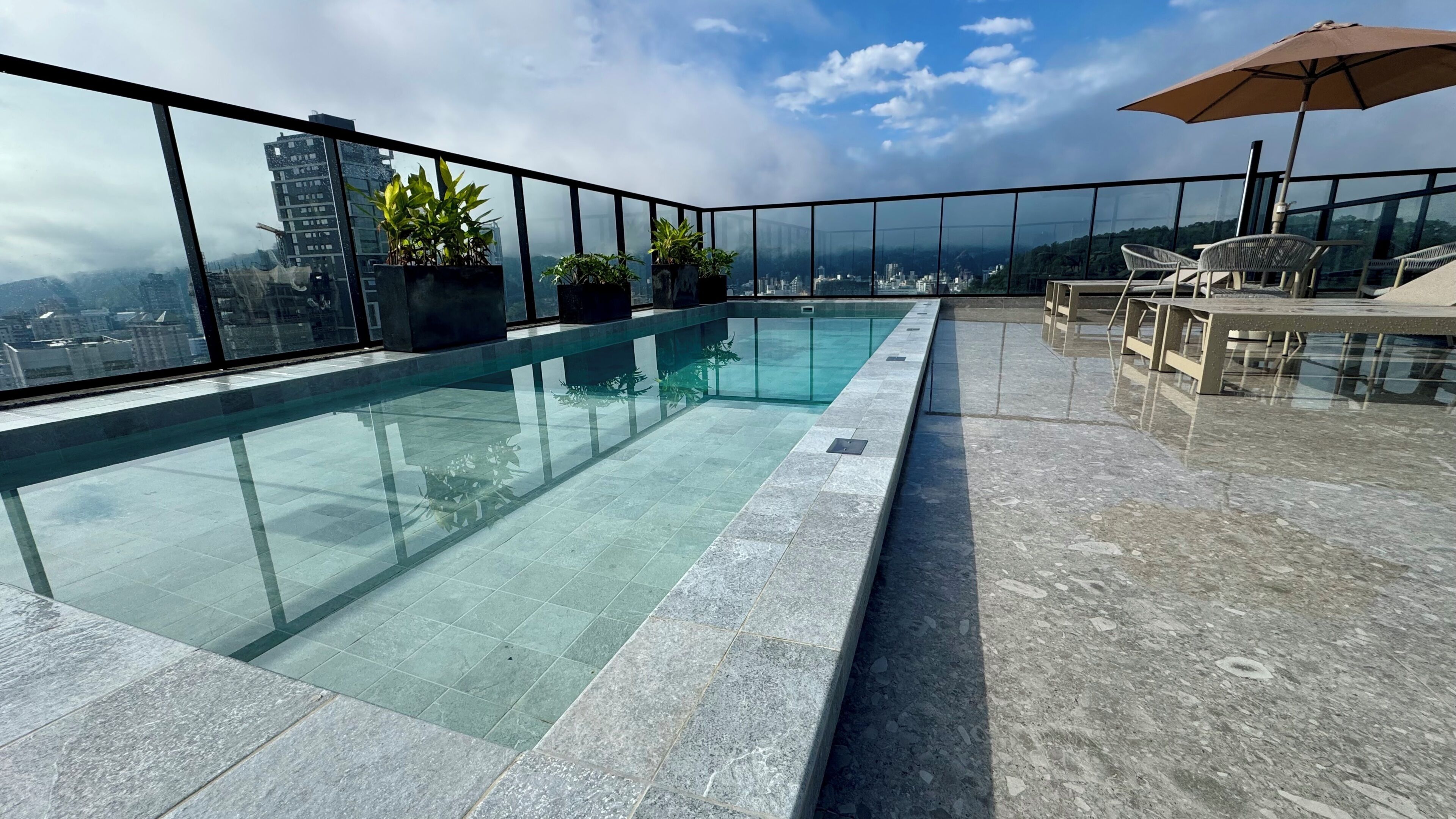 A rooftop infinity pool with clear glass edges, surrounded by potted plants, and overlooking a cityscape.