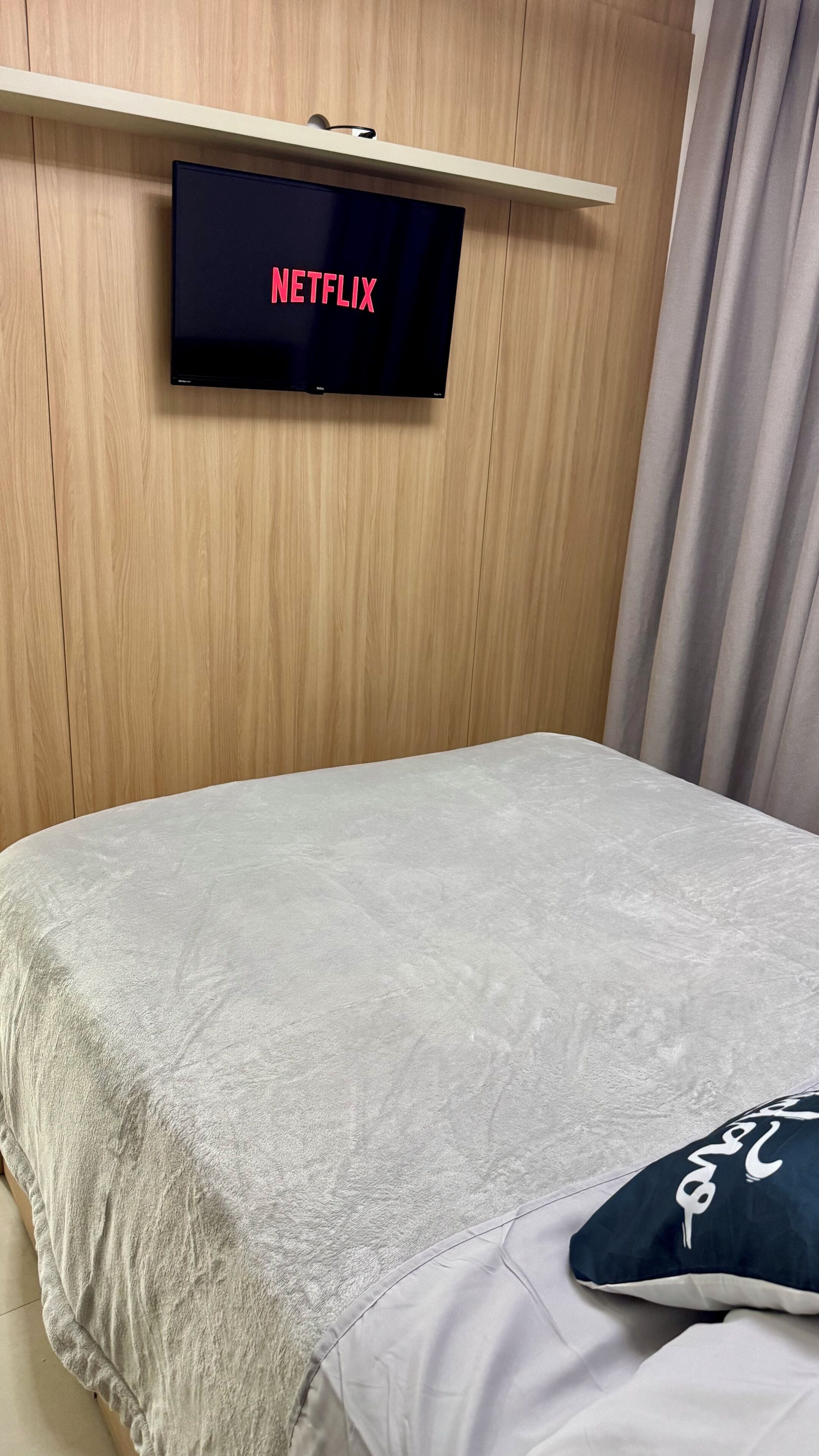 A room with a bed, a wall-mounted television displaying 'NETFLIX,' and a curtain.