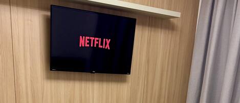 A room with a bed, a wall-mounted television displaying 'NETFLIX,' and a curtain.
