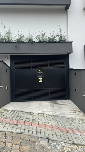 Una porta de garatge negra amb un senyal de seguretat, una entrada pavimentada amb rajoles vermelles i grises, i una paret blanca amb verdura a la part superior.