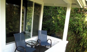 Un área de estar al aire libre con techo, con dos sillas y una mesa, rodeada de ventanales y vista a abundante follaje.