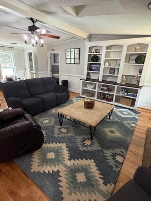 Ein Wohnzimmer mit einem dunkelgrauen Sofa, einem Couchtisch und einem gemusterten Teppich. Eine weiße Regaleinheit enthält verschiedene dekorative Gegenstände.