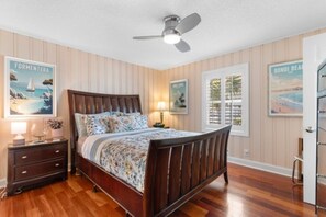 A bedroom with a wooden bed, a nightstand, a ceiling fan, and wall art depicting beaches. - The Gatsby House - Ocean View - Steps to Beach - 8BR 6BA (Murrells Inlet)
