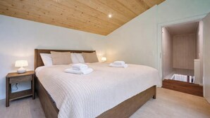 Ein Schlafzimmer mit Holzbalkendecke, einem Bett mit weißer Bettwäsche, zwei Nachttischlampen und einem ordentlich bezogenen Bett mit gefalteten Handtüchern.