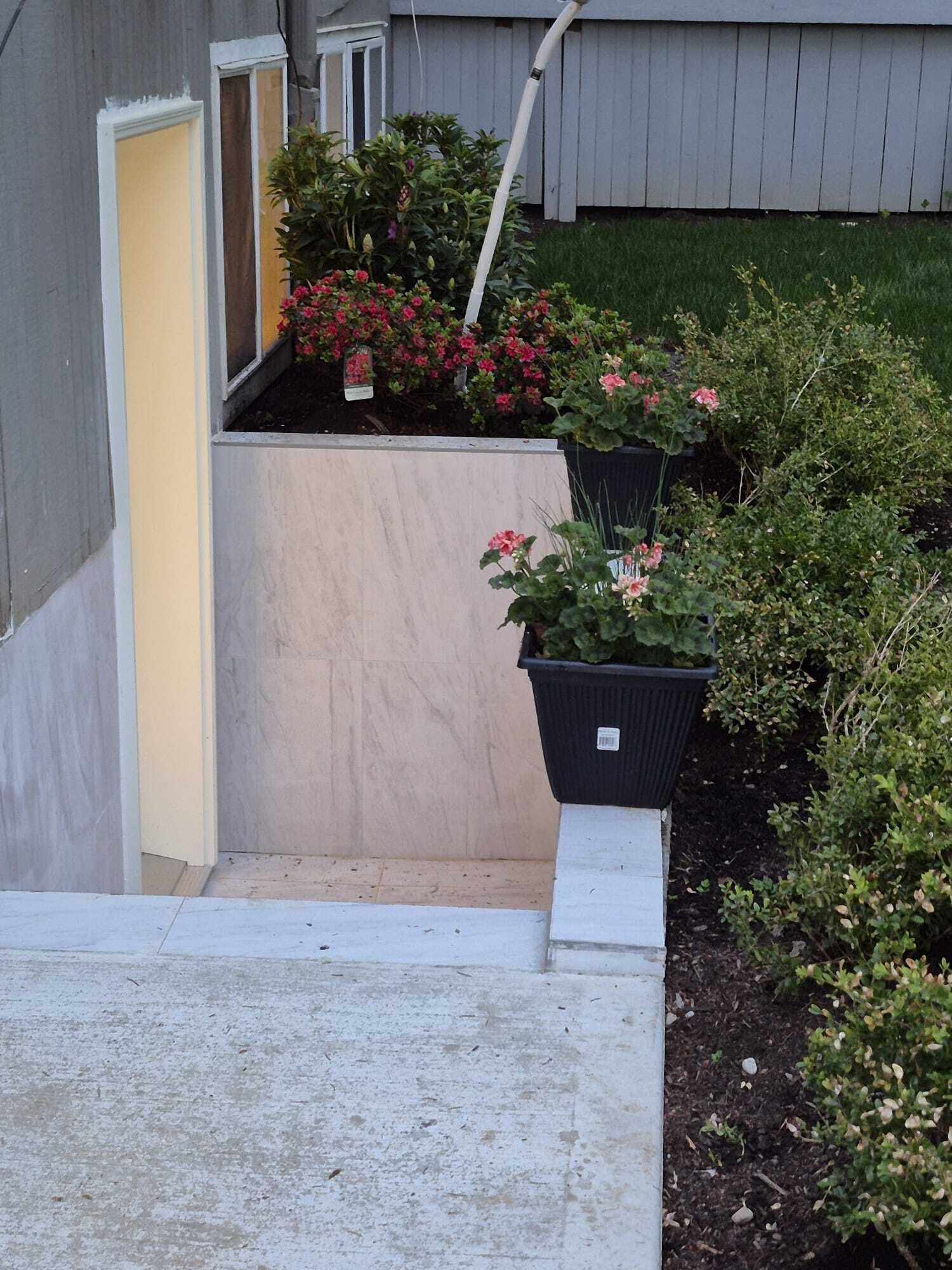 A residential entryway with a concrete step leading to a door, a small garden area with potted plants, and a wooden fence in the background.