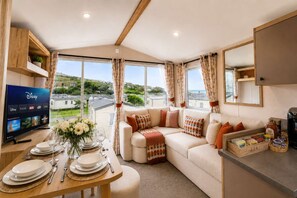 A cozy living room with a sofa, a television, and a dining area with a table set for two. - Sandy Toes New 2026 Stylish 2Bed Steps from Beach (Challaborough)