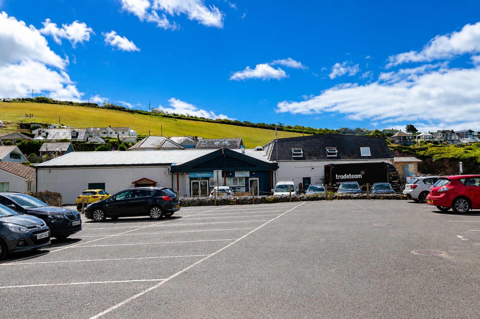 A parking lot with several cars, a building with a sign that reads 'tradetean,' and a hilly landscape in the background.