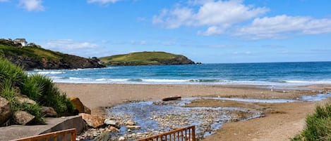 Una playa costera con un litoral rocoso, una colina cubierta de hierba al fondo y un cielo azul despejado.