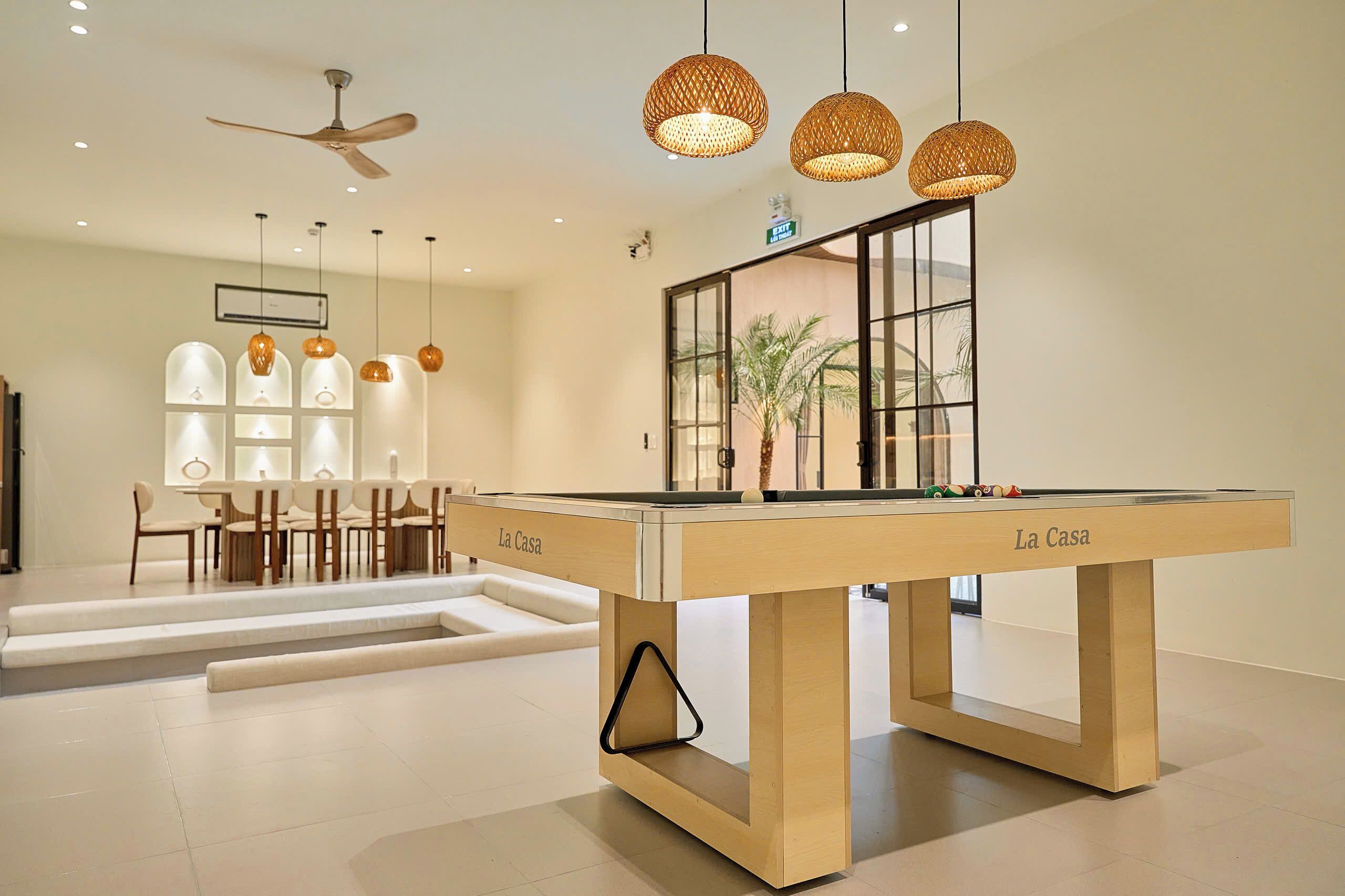 Uma mesa de bilhar moderna em um ambiente bem iluminado, com ventilador de teto, luminárias pendentes e vista para o exterior através de amplas janelas.