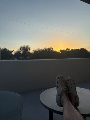 A person seated with their feet on a table, overlooking a sunset with trees in the background.