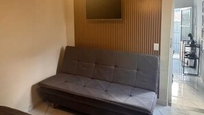 A room with a sofa, a television mounted on a wooden panel wall, and a tiled floor. - Estúdio 4 - Vila Granados em Embu das Artes (Embu das Artes)