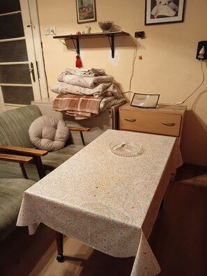 Ein Zimmer mit einem Tisch, der mit einer gemusterten Tischdecke bedeckt ist, einem grünen Sessel, einem Holzstuhl, einem kleinen Nachttisch mit einem Taschentuchhalter und einem an der Wand befestigten Regal mit verschiedenen Gegenständen.