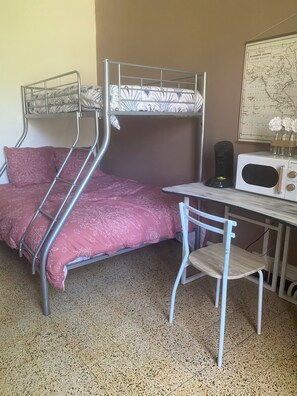 Een stapelbed met een roze beddegoed met een patroon, een witte stoel, een magnetron en een kaart aan de muur.
