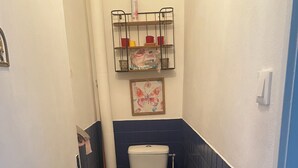 A small bathroom with a toilet, a shelf above the toilet, and a drain in the floor. - Vrbo Property (Caudiès-de-Fenouillèdes)
