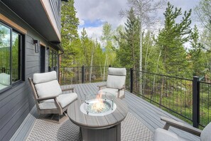 A deck with a fire pit, two chairs, and a railing overlooking a forest. - New 5 Bedroom Luxury Mountain Retreat w/Hot Tub, Private Wooded Lot - Sleeps 8 (Silverthorne)