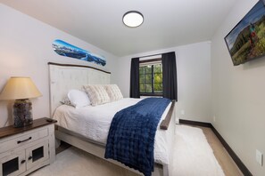 A bedroom with a bed, a nightstand, a lamp, a window with curtains, and a wall-mounted picture. - New 5 Bedroom Luxury Mountain Retreat w/Hot Tub, Private Wooded Lot - Sleeps 8 (Silverthorne)